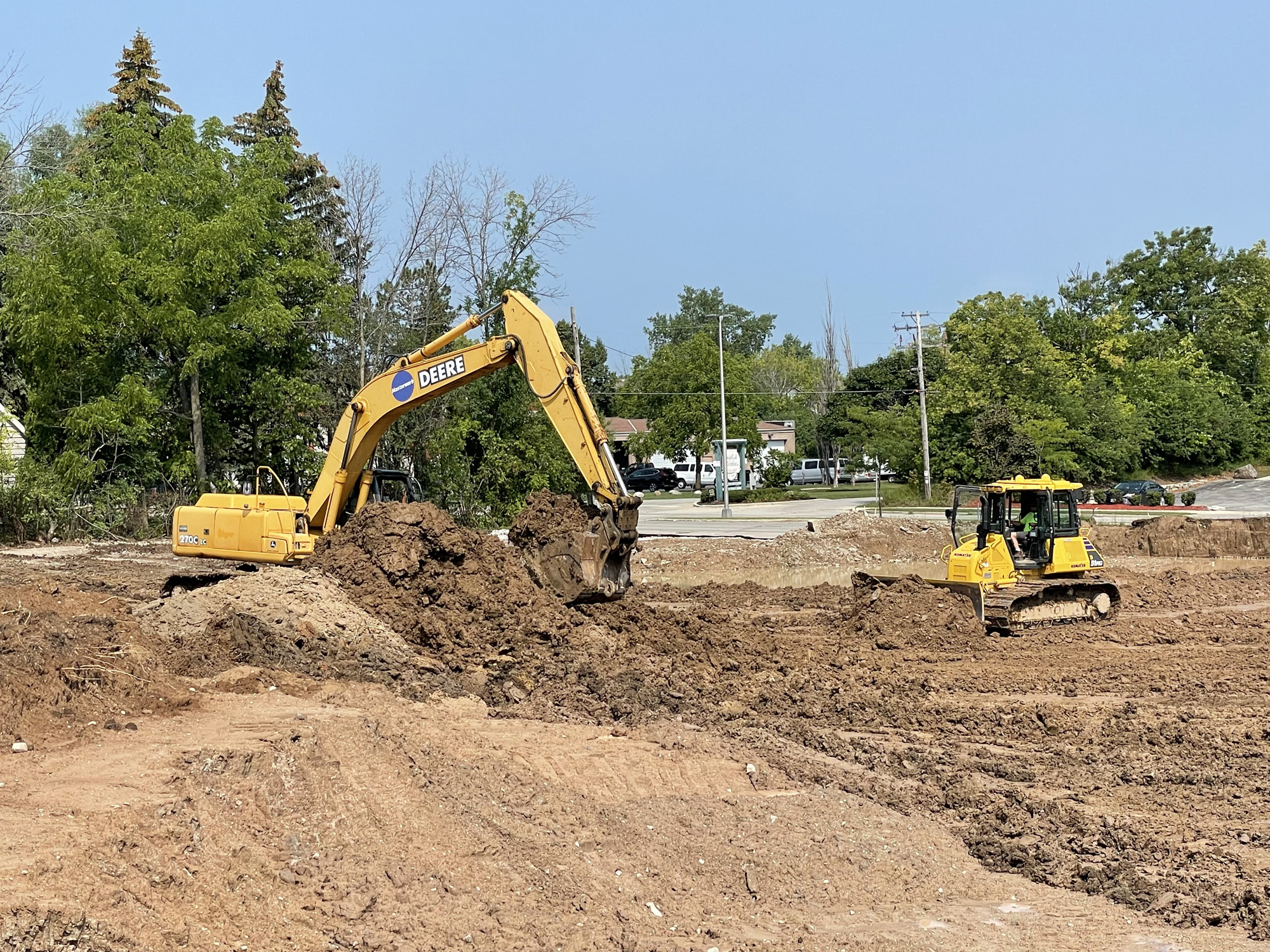 A yellow excavator and bulldozer performing land clearing operations on a sunny day in Mequon, with trees and a street in the background.